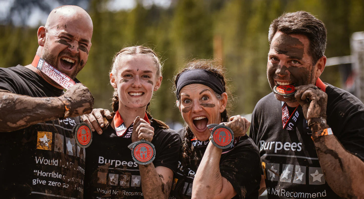Four smiling Spartan OCR participants wearing black Burpees branded athletic t-shirts and face paint, holding up their medals after an outdoor obstacle race.